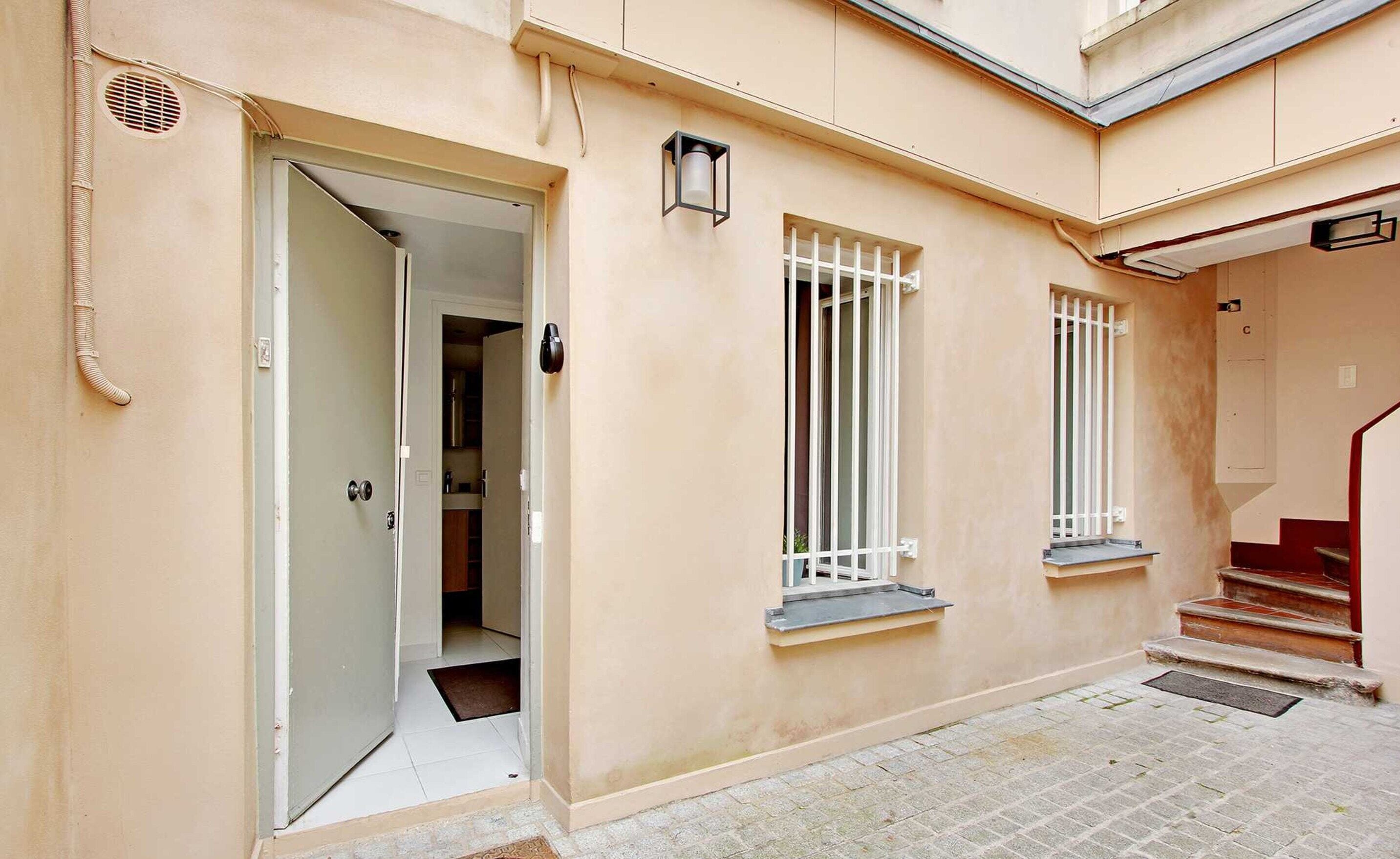 Fachada de un edificio con una puerta que da a una habitación, una ventana con celosía blanca y una escalera con barandilla roja.