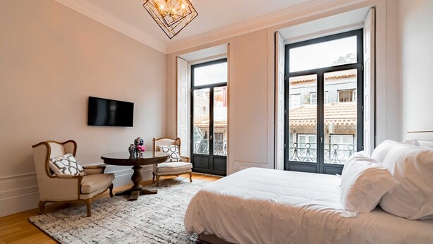 Un dormitorio con una cama grande, un televisor montado en la pared, una mesa de comedor con sillas y un balcón con vistas a edificios.