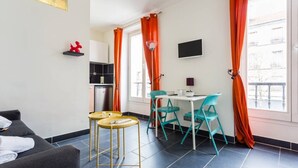 A modern living room with a sofa, a small dining table with chairs, a kitchen area, and large windows with orange curtains. - Magnificent Studio - 2P - Pantin (Pantin)