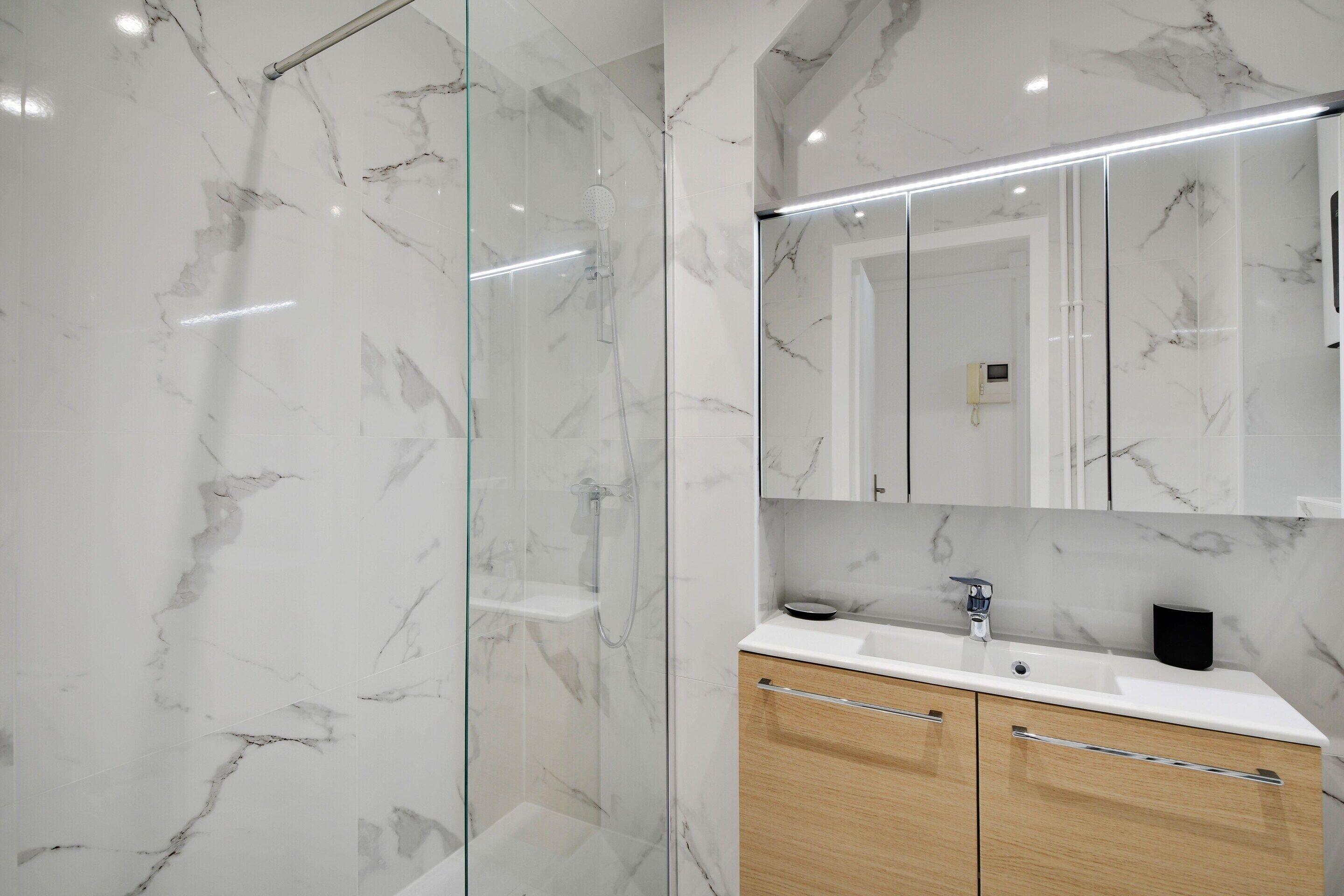 A modern bathroom with a glass shower enclosure, a marble-tiled wall, a wooden vanity with a sink, and a large mirror above the vanity.