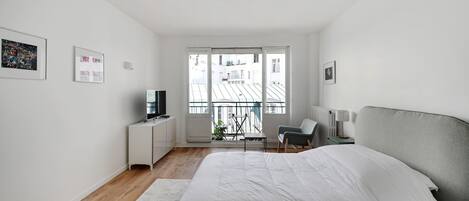 Un dormitorio moderno con una cama grande, un escritorio con televisor, una silla y un balcón con vistas a edificios.