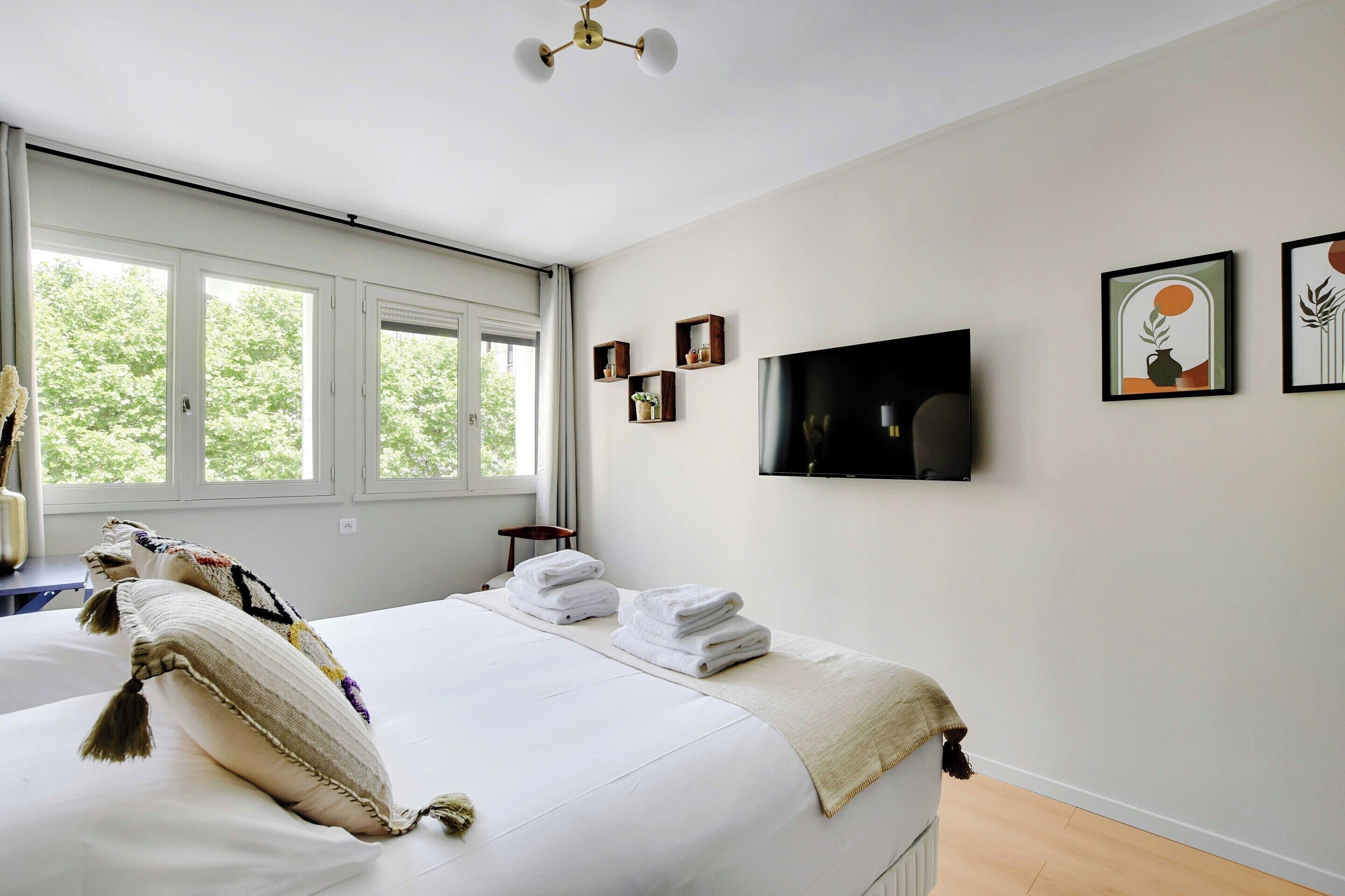 A hotel room with a large bed, a TV mounted on the wall, and a window with a view of trees.