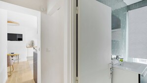 A modern interior with a white door leading to a bathroom with a large sink and a window. A television is mounted on the wall, and a small table with a chair is visible in the adjacent room. - Charming Studio for 2 People - Bastille (Paris)