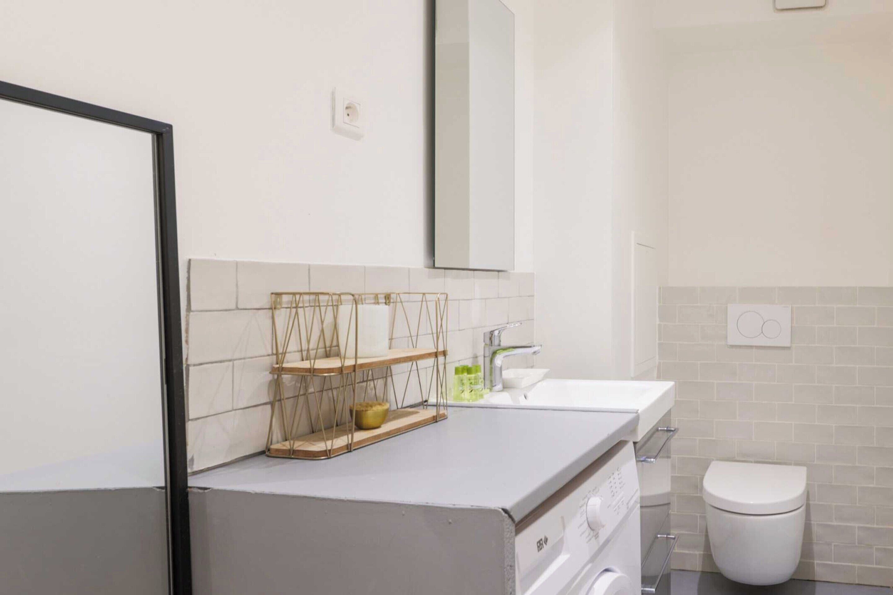 A modern bathroom with a white sink, a toilet, and a mirror. There are shelves with toiletries and a towel rack.