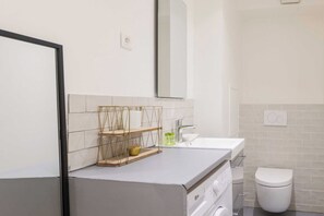 A modern bathroom with a white sink, a toilet, and a mirror. There are shelves with toiletries and a towel rack. - Charming Studio Apartment for 2 - Bastille (Paris)
