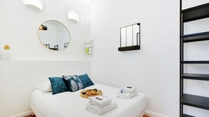 Moderni hotellihuone, jossa on sänky, pyöreä peili, pieni hylly henkilökohtaisille hygieniatarvikkeille ja musta metallinen hyllykkö.