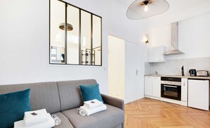 A modern, well-lit living room with a grey sofa, a large window, and a kitchen area with white cabinets and appliances.