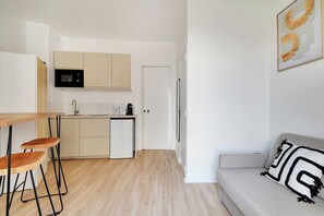 A compact kitchen with a microwave, sink, and a small refrigerator. There is a wooden dining table with two chairs and a grey sofa with a patterned pillow. - Pleasant Studio - 2P - Gateway to Paris/malakoff (Malakoff)