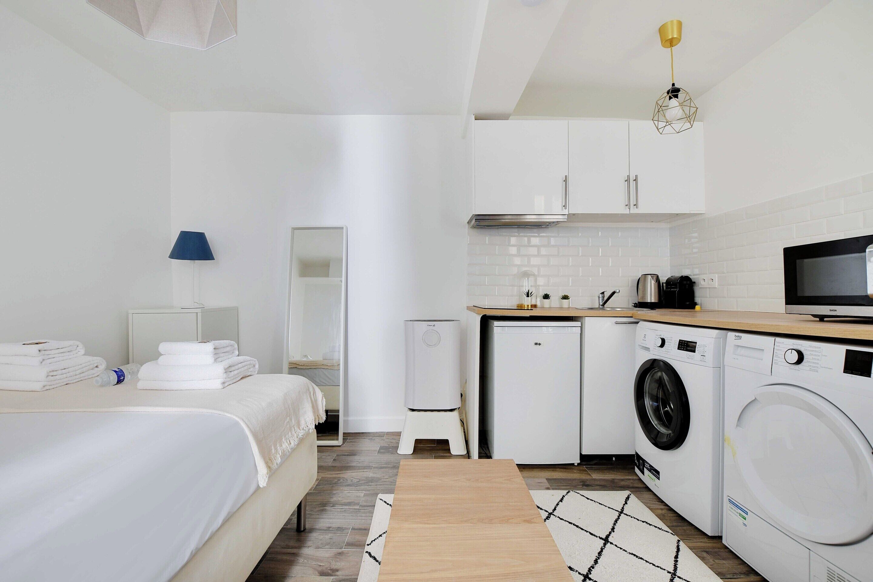 Een moderne hotelkamer met een bed, nachtkastje, lamp, wasmachine, droger en kitchenette.