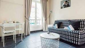 Ein Zimmer mit einer gemusterten Couch, einem kleinen runden Tisch, einem weißen Stuhl und einem Fenster mit Vorhängen.
