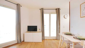 Ein helles Zimmer mit Holzboden, einem weißen TV-Schrank, einem Esstisch mit Stühlen und Blick durch eine Glastür.
