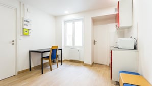 A small, well-lit room with a wooden floor, a table with a chair, a window, a white radiator, and a small kitchenette with a microwave. - Elegant Studio - 2P - Malakoff (Malakoff)