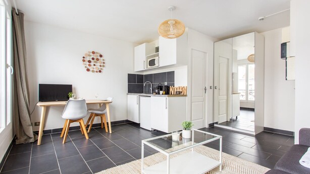 Een moderne woonkamer met een glazen salontafel, een eethoek met een witte tafel en stoelen, een keuken met witte kastjes en een zwart aanrecht, en een groot raam met gordijnen.