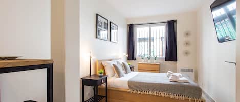 A bedroom with a bed, bedside tables, a window with curtains, a wall-mounted TV, and a small plant.