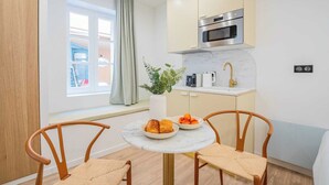 Cocina compacta con horno integrado, una pequeña encimera y una ventana con cortinas. Hay una mesa redonda con dos sillas y un jarrón con una planta encima.