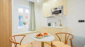 Cocina compacta con microondas, horno y lavaplatos. Hay una pequeña mesa redonda con sillas y una ventana con cortinas.