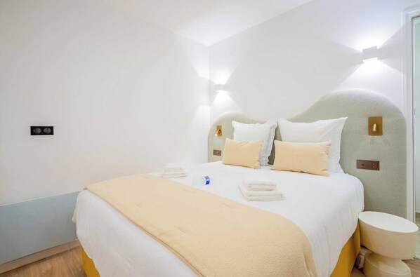A neatly made bed with white and beige bedding, a bedside table with a lamp, and a wall-mounted light fixture.