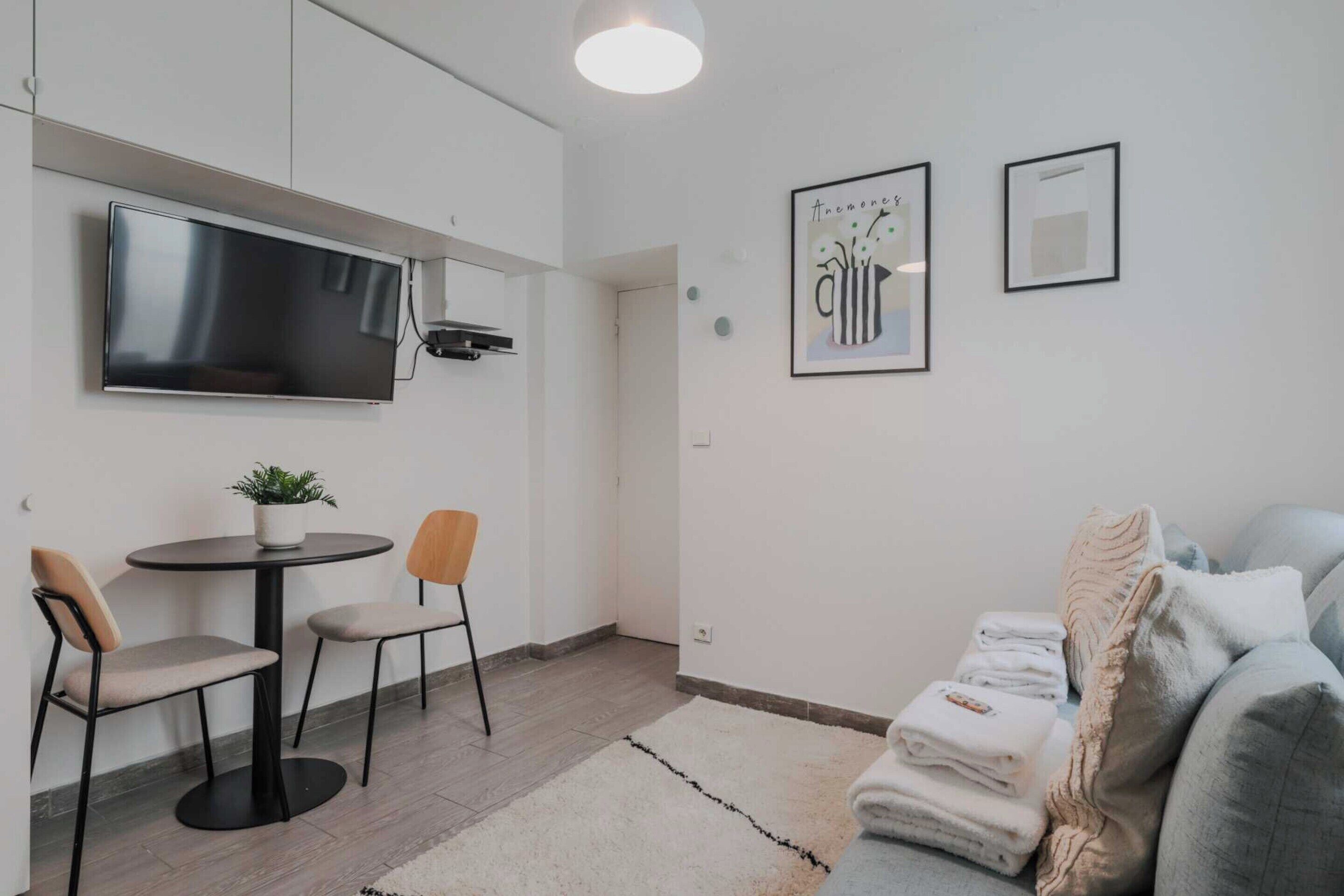 A modern living room with a flat-screen TV, a round table with two chairs, a grey sofa, and a few framed artworks on the wall.