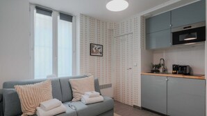 Een moderne woonkamer met een grijze bank, twee witte kussens en een beige plaid. Een keuken met grijze kasten en een zwarte oven is zichtbaar.