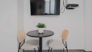 Een vergaderruimte met een flatscreen televisie aan de muur, een tafeltje met een bloempot en twee stoelen met lichte houten frames en grijze kussens.