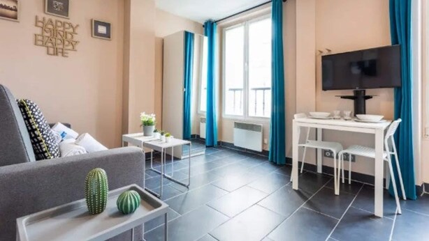 Un salon moderne avec un canapé, une table basse ornée d’un cactus, un coin repas avec un téléviseur et une grande fenêtre avec des rideaux.