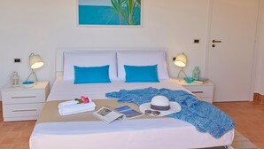A neatly made bed with blue pillows, a white headboard, and a framed painting above it. - Villa Rosa dei Venti, Scopello (CASTELLAMMARE DEL GOLFO)