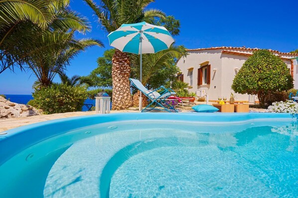 A poolside area with a clear blue pool, lounge chairs, a stone pillar, and a building with a tiled roof. - Villa Ginestra, Scopello (CASTELLAMMARE DEL GOLFO)
