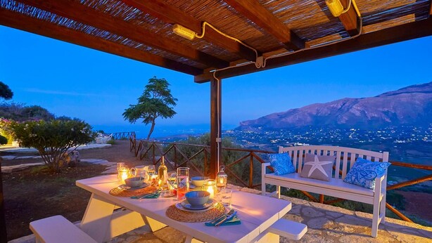 An outdoor dining area with a table set for two, a bench, and a view of a cityscape at dusk. - Villa Delfino, Scopello (CASTELLAMMARE DEL GOLFO)