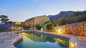 A pool with stone tiles, a stone wall, and a building with lights. - Villa Delfino, Scopello (CASTELLAMMARE DEL GOLFO)