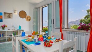 A bright room with a dining table set for breakfast, a bookshelf, and a view of the outdoors. - Villa Delfino, Scopello (CASTELLAMMARE DEL GOLFO)