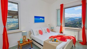 A bedroom with a bed, a view of the countryside, a window with curtains, and a small table with a lamp and flowers. - Villa Delfino, Scopello (CASTELLAMMARE DEL GOLFO)