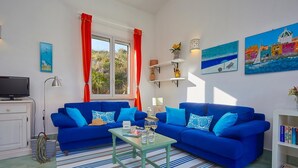 A living room with two blue sofas, a striped rug, a small table with drinks, a TV, and a bookshelf. - Villa Delfino, Scopello (CASTELLAMMARE DEL GOLFO)
