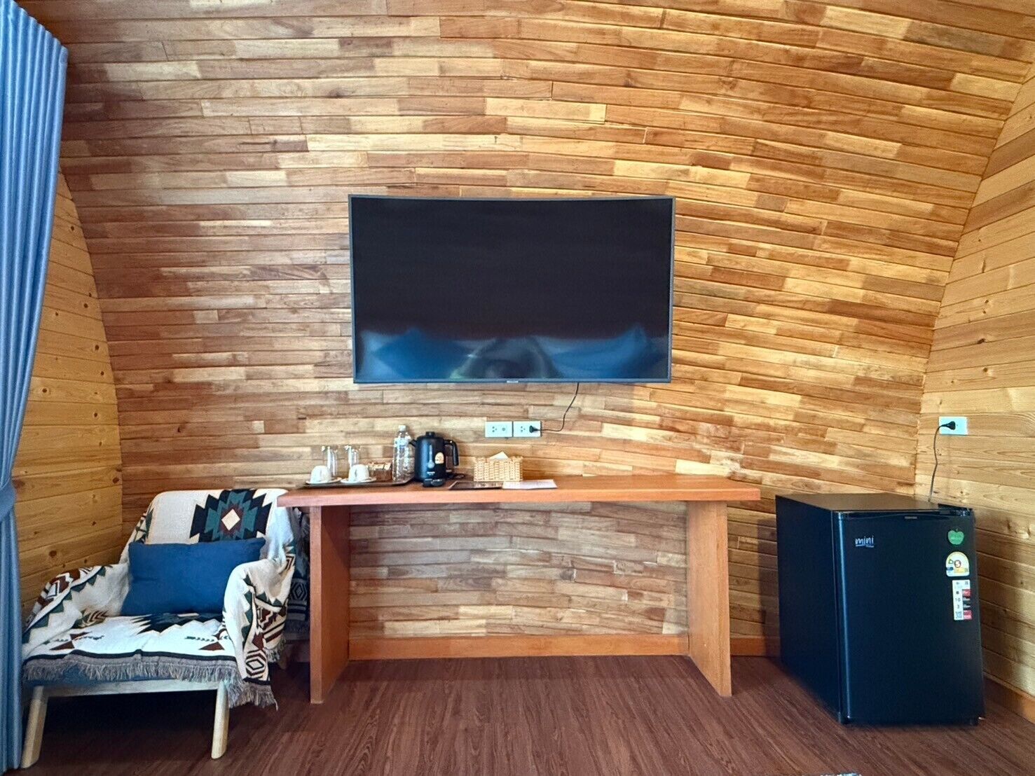 Una habitación acogedora con un muro de madera, un televisor de pantalla plana sobre un escritorio de madera, un refrigerador negro pequeño y una silla estampada.