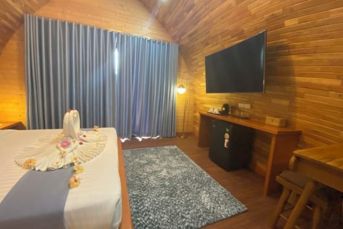 A cozy room with a bed, a desk, a TV mounted on the wall, and a large window with curtains.
