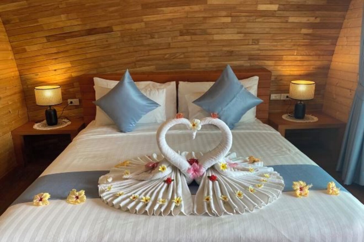 A hotel room with a wooden ceiling, two nightstands with lamps, a bed with white bedding, blue pillows, and a heart-shaped towel arrangement on the bed.