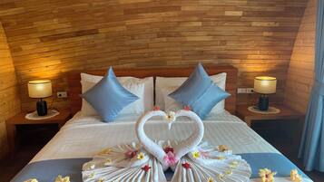 Habitación de hotel con techo de madera, cama con ropa blanca, dos almohadas azules y una decoración con toallas en forma de corazón y flores.