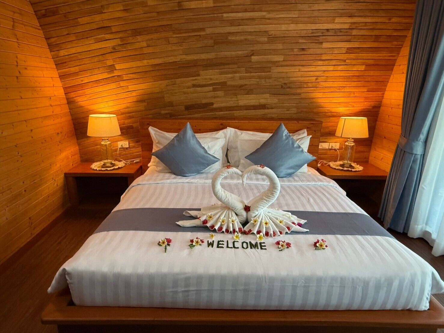 Ein Hotelzimmer mit Holzbalkendecke, einem Bett mit der Aufschrift 'WELCOME', Kissen und zwei Nachttischlampen.