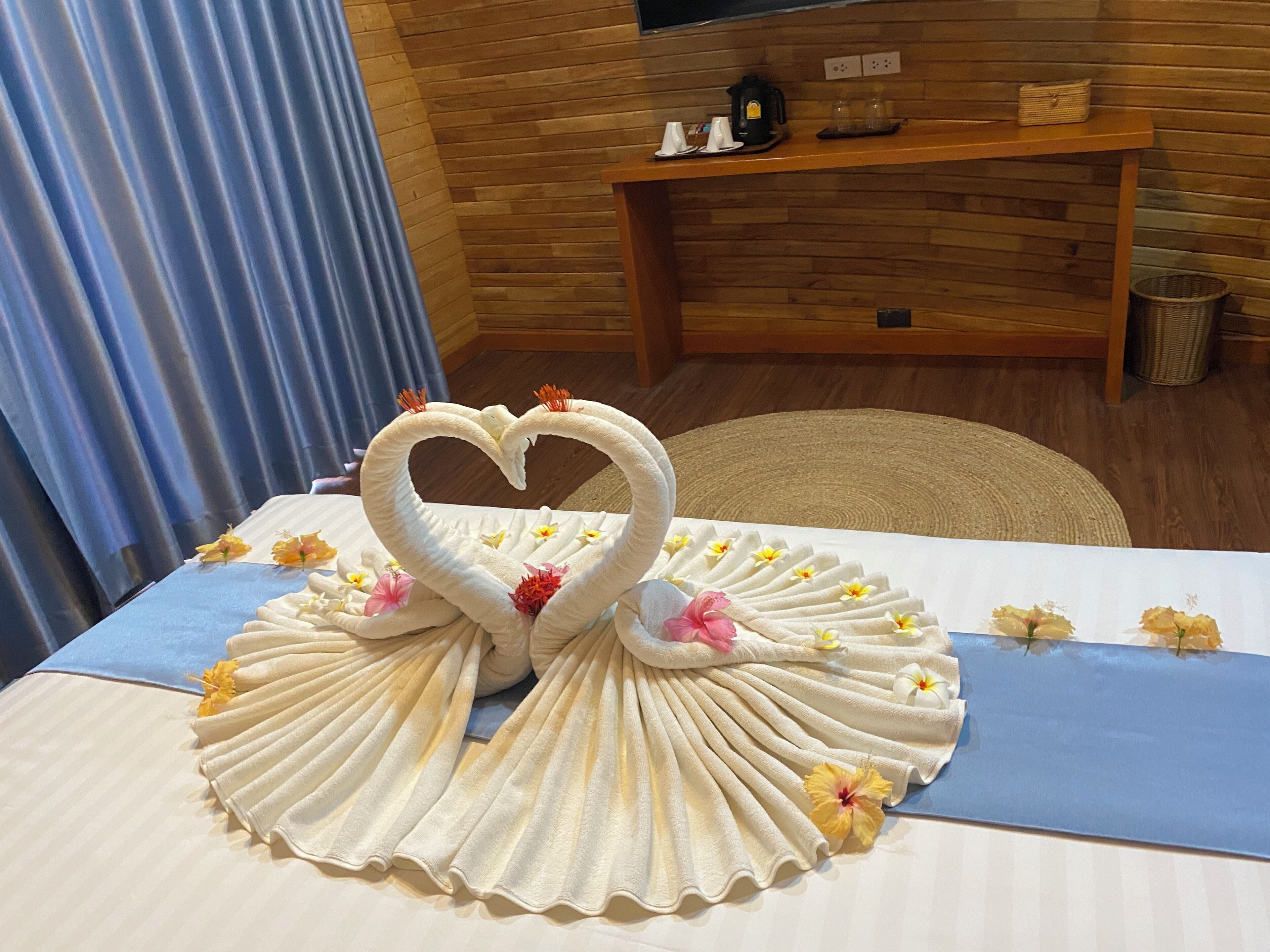 Una habitación con pared de madera, cortina azul, mesa con un arreglo de toallas en forma de cisnes y un estante con objetos.