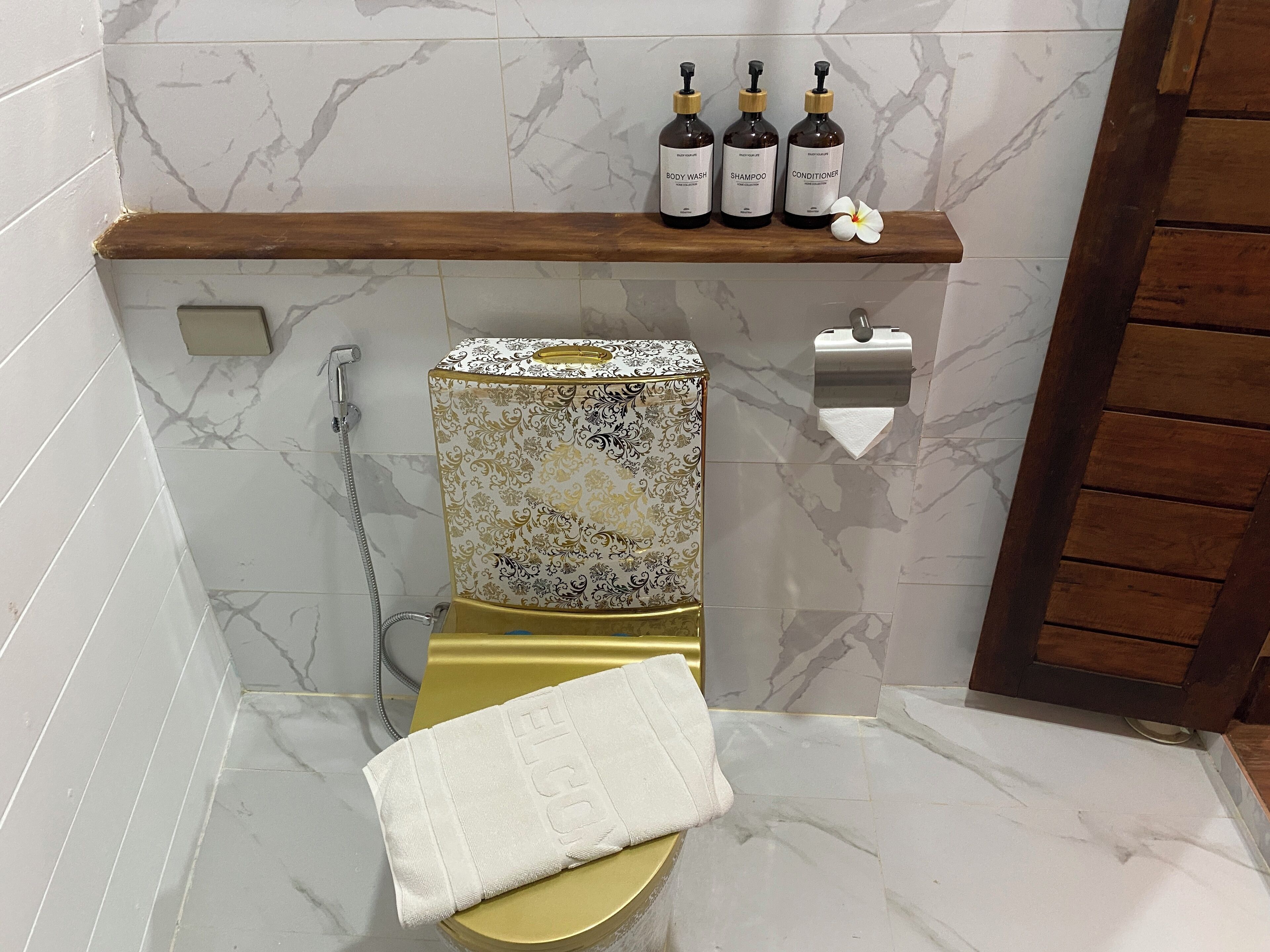 A bathroom with a gold toilet, a marble wall, and a shelf with three bottles of toiletries.