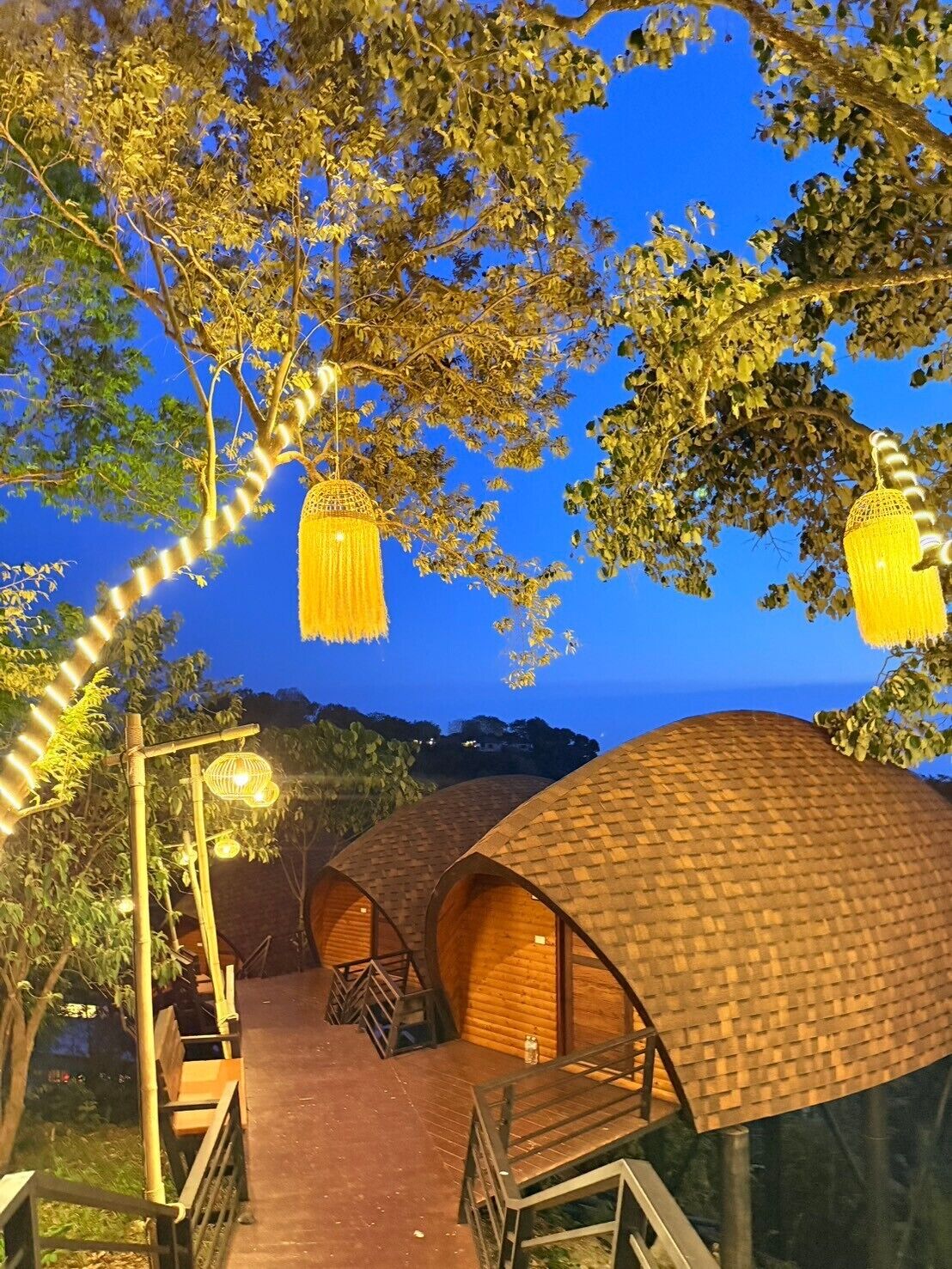 A row of wooden dome-shaped cabins with a wooden walkway leading to them, surrounded by trees and string lights.