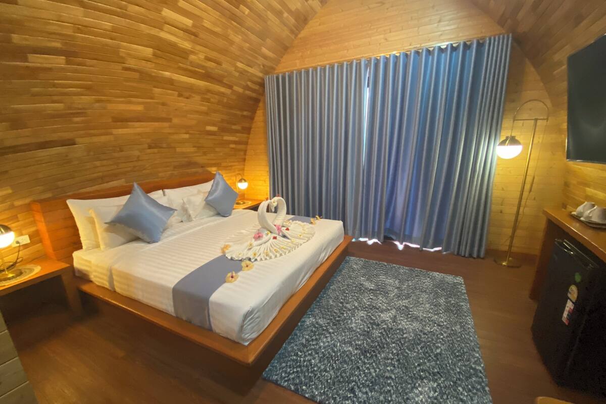 Un dormitorio acogedor con techo de madera, una cama bien tendida con almohadas, una cortina azul y un escritorio pequeño con una lámpara.