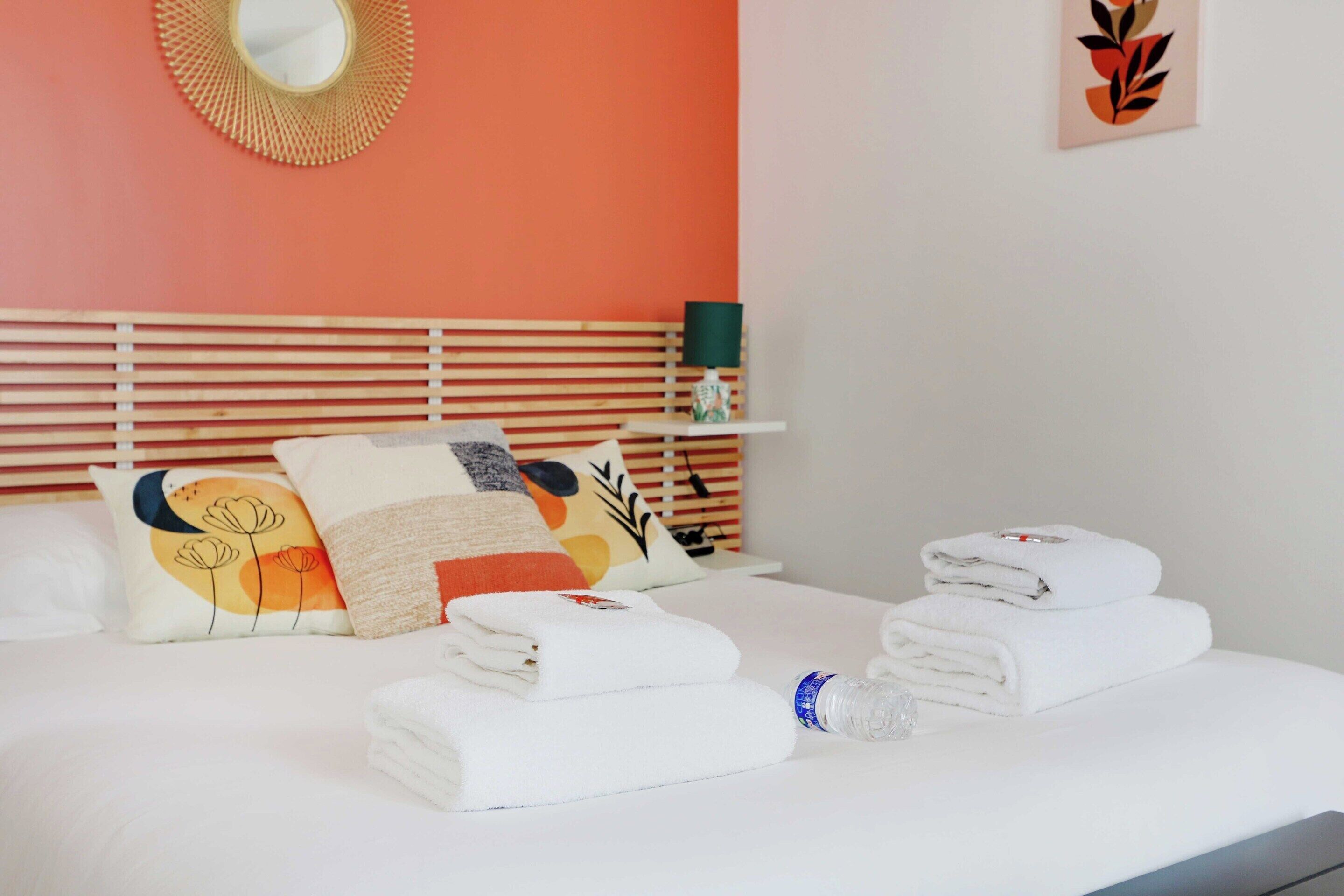 A neatly made bed with white linens, a decorative wall mirror, and a small shelf with a lamp and a vase.