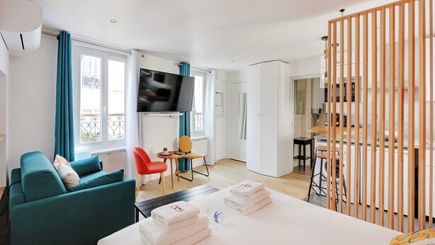 A modern hotel room with a sofa, a dining area, a TV, and a kitchen in the background. - Stunning Studio for 2 People - Sentier (Paris)
