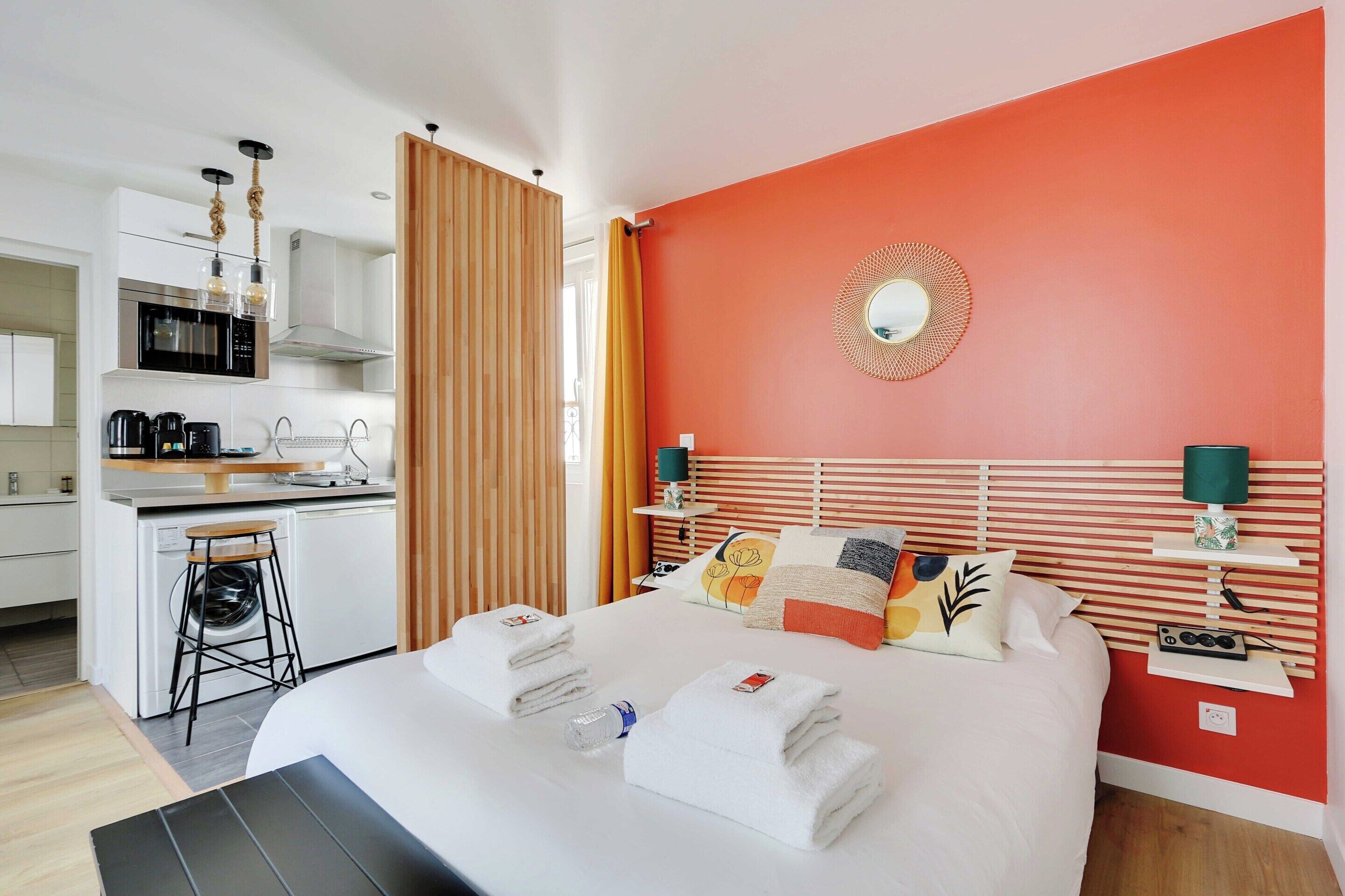 A hotel room with a bed, a nightstand, a mirror, a window with curtains, a kitchen area, and a bathroom.