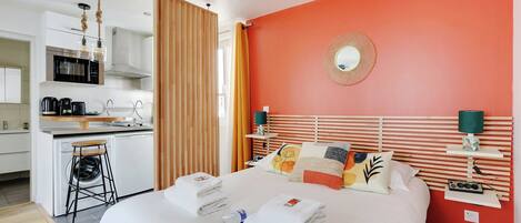 A hotel room with a bed, a nightstand, a mirror, a window with curtains, a kitchen area, and a bathroom.
