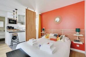 A hotel room with a bed, a nightstand, a mirror, a window with curtains, a kitchen area, and a bathroom. - Stunning Studio for 2 People - Sentier (Paris)