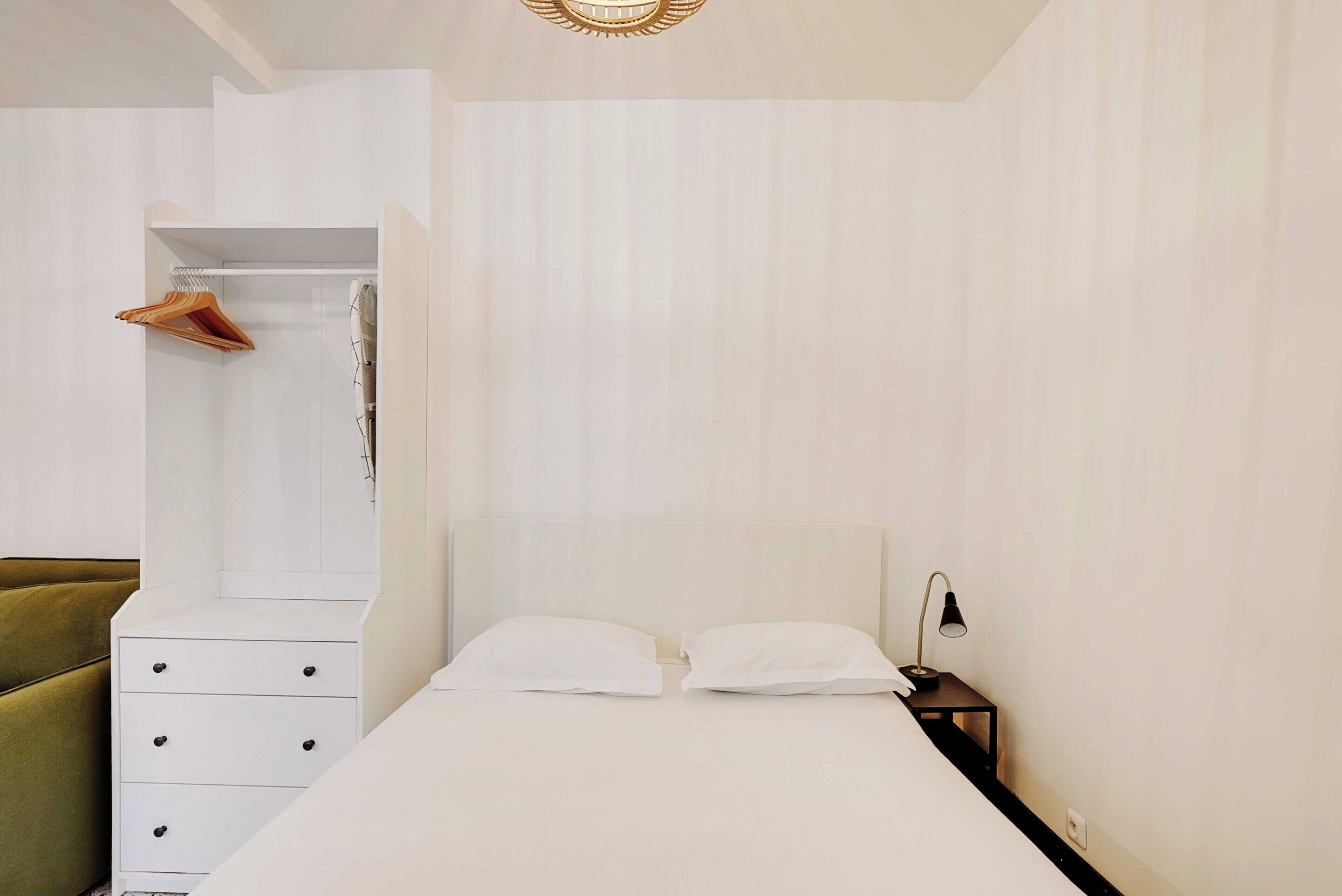 A minimalist bedroom with a bed, a bedside table with a lamp, a wardrobe, and a green chair.
