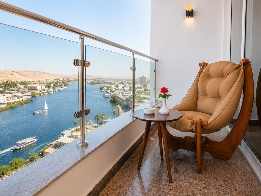 A balcony with a view of a river, a chair, a small table with a vase and a bowl, and a wall-mounted light fixture.
