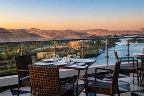 Un espace repas avec des tables préparées pour deux personnes, offrant une vue sur une rivière et des montagnes.