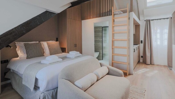 Un dormitorio moderno con cama estilo loft, armario empotrado, escalera al nivel superior y un amplio ventanal que proporciona luz natural.